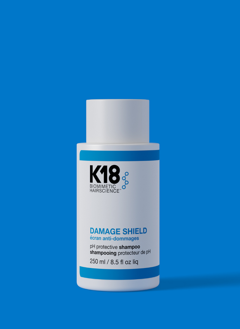 A bottle of DAMAGE SHIELD protective shampoo by K18, with a white and blue label, indicating the size of 8.5 fl oz.