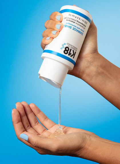Hand holding a K18 product with clear liquid being dispensed against a blue background